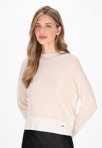 Helles beiges Strickpullover mit langen Ärmeln, geripptem Saum und dezenter Logodetail; glatte Textur und lockere Passform.