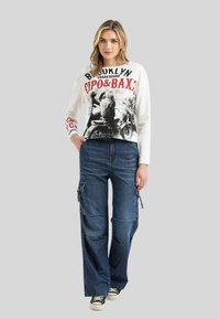 Langarmhemd in Weiß mit Grafikdruck, kombiniert mit blauen Cargo-Jeans mit Seitentaschen und lässiger Passform, abgerundet mit schwarzen Sneakers.