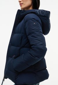 Femme portant une doudoune bleu marine avec une capuche et un patch logo rond sur le haut de la manche, mains dans les poches, vue de profil.