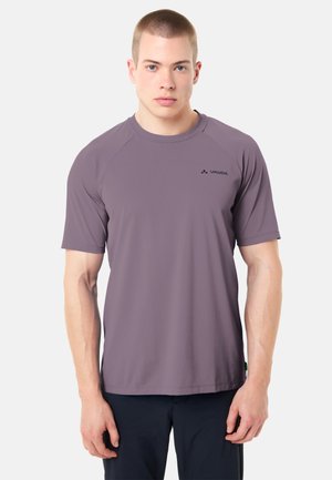 Junger Mann mit kurzen Haaren, der ein schlichtes, lila, kurzärmeliges Sport-T-Shirt und dunkle Hosen trägt, schaut nach vorne vor einem weißen Hintergrund.