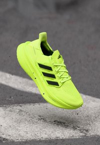 Fel geelgroene hardloopschoen met zwarte strepen, zwevend boven asfalt met een witte wegbelijning.