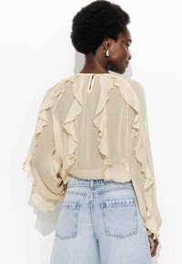 Blouse beige avec des détails froncés le long des manches et du dos, dotée d'une fermeture en forme de clé. Assortie à un jean taille haute bleu clair.