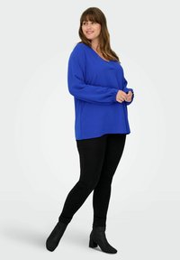 Blaues, langärmeliges Oberteil mit V-Ausschnitt und gerafften Manschetten, kombiniert mit schwarzen Leggings und Knöchelstiefeln. Glatte Textur; lässige Passform.