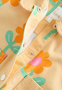 Tissu jaune avec des fleurs orange et rose ainsi que des feuilles vertes, avec des boutons-pression et un col partiellement ouvert près d'une étiquette blanche.