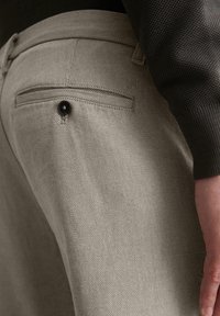 Beige strukturierte Hose mit einer Gesäßtasche und schwarzem Knopfdetail. Der Stoff hat ein gewebtes Design, eine gedämpfte Farbe und eine taillierte Passform.