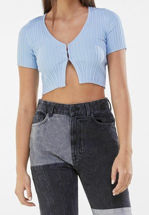 Kobieta w jasnoniebieskim prążkowanym krótkim kardiganie zapinanym na dwa hafciki oraz w czarno-szarych dżinsach z łatami.