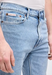 Jeans in denim azzurro chiaro indossati da una persona, con un'etichetta Calvin Klein sulla piccola tasca frontale vicino alla vita.