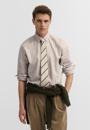 Junger Mann, der ein hell gestreiftes Hemd mit Knöpfen, eine diagonal gestreifte Krawatte, khakifarbene Cordhosen und einen dunklen Pullover, der um die Taille gebunden ist, trägt.