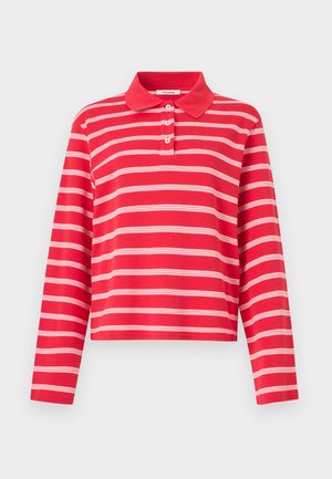 Femme portant un polo à manches longues rayé rouge et blanc, un pantalon beige à jambes larges et des sandales à talons ouvertes noires, posant sur fond blanc.