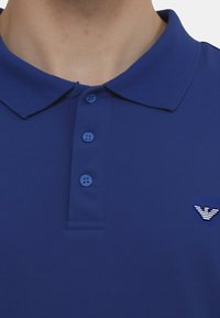 Polo shirt blu royal con colletto a coste, patta con tre bottoni e logo ricamato che presenta strisce orizzontali. Texture liscia.
