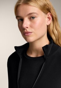 Čierna bunda s vysokým golierom na zips a hladkým povrchom, v kombinácii s čiernym tričkom s okrúhlym výstrihom. Detailný záber na golier a detaily výstrihu.