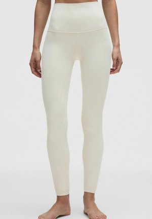 Hoch taillierte, cremefarbene Leggings an einer stehenden Person, bis zu den Knöcheln gekürzt, mit nackten Füßen vor einem einfarbigen Hintergrund.
