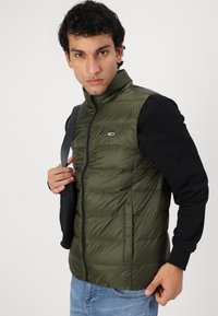 Maslinasto zeleni puffer prsluk s visokim ovratnikom, prednjim zatvaračem i horizontalnim šavovima. Crne duge rukave s detaljem na ramenu.