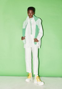 White oversized shirt over a striped green top, paired with white trousers and chunky white sneakers with yellow laces. Background is green.