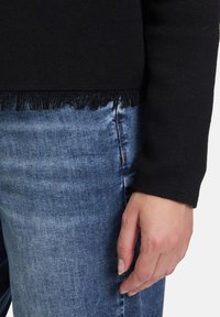 Černý pletený svetr s třásněmi na spodním lemu, doplněný modrými džíny. Záběr zblízka ruky odpočívající na džínách.