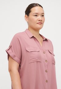 Chemise courte à manches courtes de couleur rose pâle en tissu doux, avec un col en V, un bouton sur le devant, et deux poches poitrine avec des boutons décoratifs.