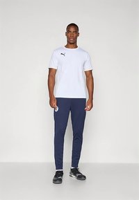 Moški stoji oblečen v belo majico Puma, temno modre hlače z logotipom in črne športne čevlje pred enobarvno svetlo ozadje.