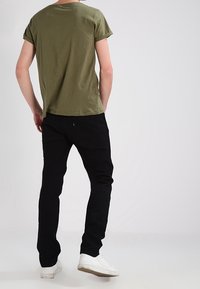 Camiseta de algodón verde oliva con mangas remangadas, combinada con pantalones negros y zapatillas blancas. Diseño sencillo y ajustado visible desde la parte de atrás.