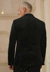 Schwarze Wildlederjacke mit tailliertem Schnitt, ausgestattet mit einem einzelnen Schlitz, strukturiertem Stoff und knöpfbaren Ärmeln, von hinten betrachtet.