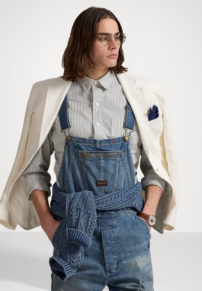 Salopette en denim avec une poche zippée et des bretelles, assortie d'une chemise rayée et d'un blazer crème. Comprend un pull bleu tricoté noué à la taille.