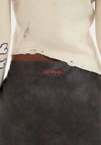 Suéter de punto en color crema con dobladillo deshilachado, combinado con una falda de cuero sintético gris oscuro que presenta el logo rojo "Ed Hardy" en la cintura.