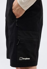 Άτομο που φοράει μαύρα σορτς Berghaus με φερμουάρ στην πλαϊνή τσέπη και το χέρι εν μέρει μέσα στην μπροστινή τσέπη, μπροστά σε απλό φόντο.