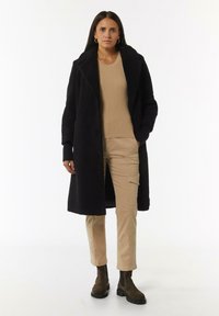 Schwarzer, strukturierter langer Mantel mit einem breiten Kragen, kombiniert mit einem beigen gerippten Oberteil und hellbeigen Cargo-Hosen. Braune Ankle Boots mit dicken Sohlen.