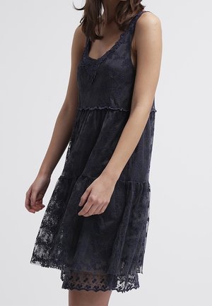 Abito senza maniche in pizzo blu navy con motivo floreale, gonna a balze e orlo smerlato indossato da una persona su uno sfondo semplice.