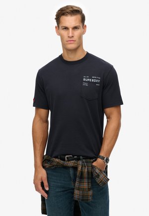 Navy blue short-sleeve t-shirt with a chest pocket featuring white text. Model wears it with a plaid shirt tied at the waist and denim jeans.