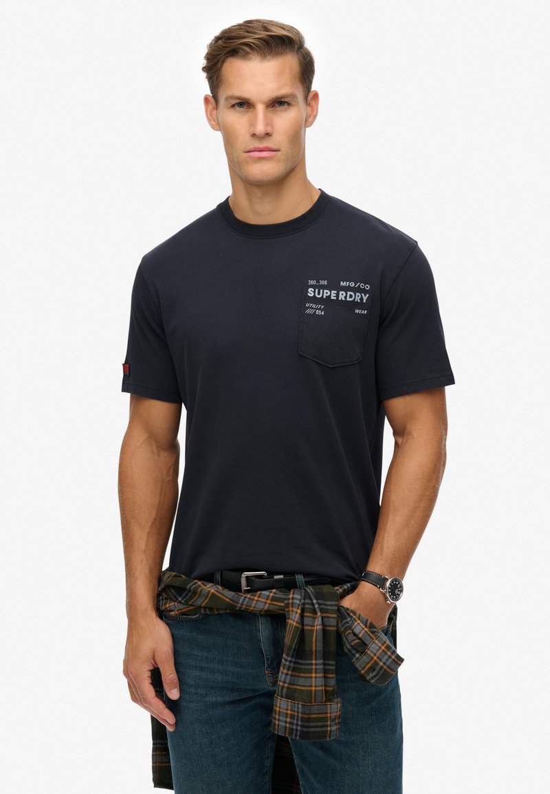 Marineblå kortærmet t-shirt med en brystlomme, der har hvid tekst. Modellen har den på sammen med en ternet skjorte knyttet om taljen og denim jeans.