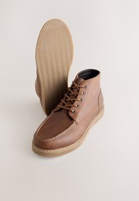 Bottes courtes en cuir marron avec lacets et détails de coutures, semelle en caoutchouc texturée à motif chevrons, et finition supérieure lisse.