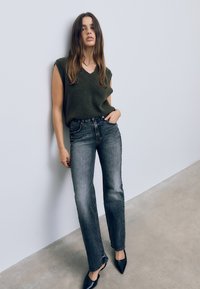 Temno zelena brezrokavnik s dizajnom v obliko črke V, v kombinaciji z visokim pasom, ravnimi bledimi črnimi kavbojkami in koničastimi črnimi petami.