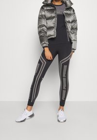 Silvrig puffjacka med hög krage, svart åtsittande topp och svarta leggings med grafiska mönster. Vita skor med svarta accenter.