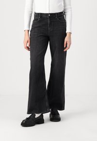 High-waisted black denim flared jeans with front pockets, faint texture, and a button closure. Paired with black loafers and a white top.