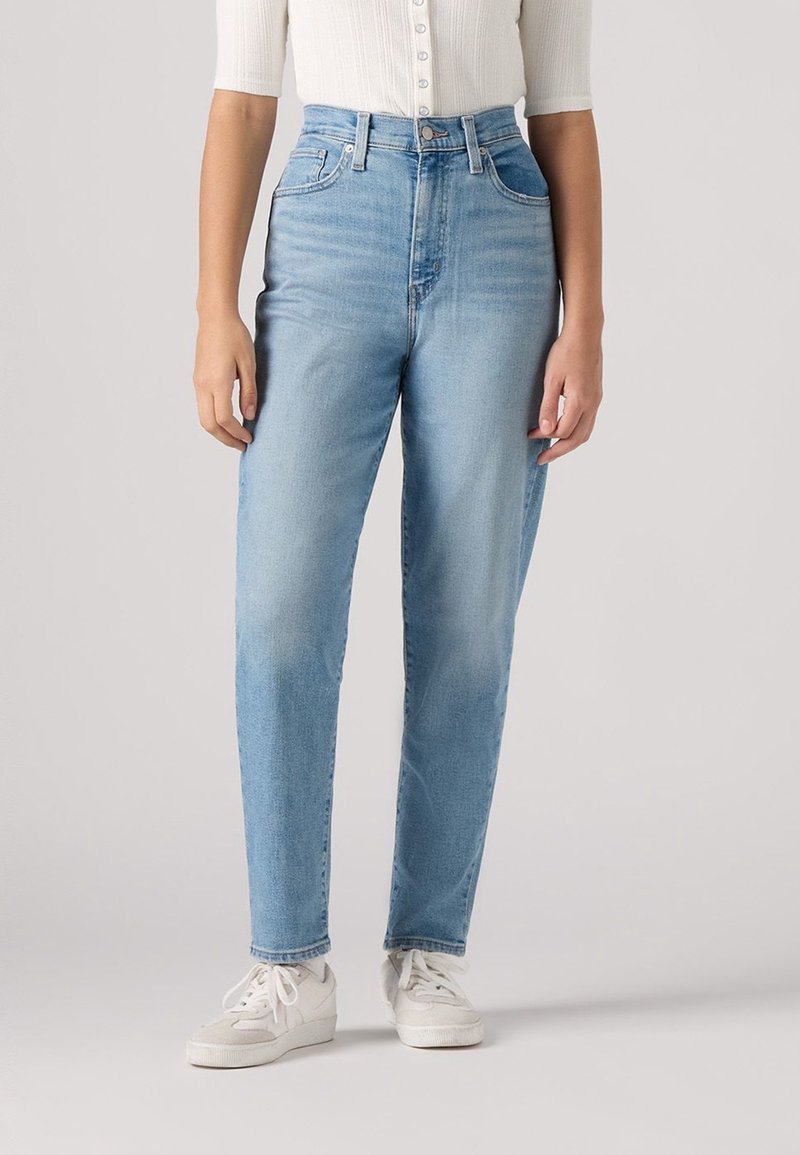 Jean taille haute bleu clair en denim, avec une coupe droite, des poches avant, et une légère décoloration, porté avec des baskets blanches.