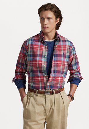 Camisa de cuadros roja y azul con botones y mangas largas, superpuesta a un suéter azul marino, combinada con pantalones de tela beige con pliegues y un cinturón marrón.
