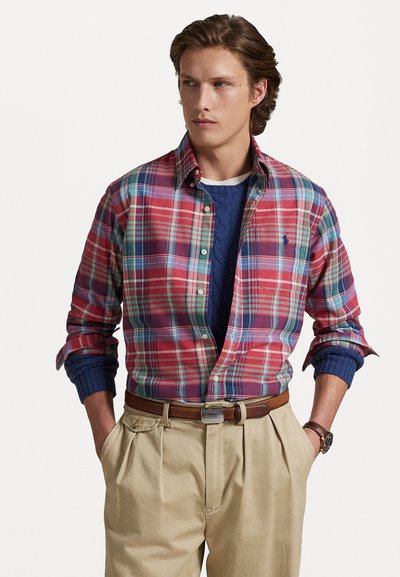 Chemise à boutons à carreaux rouges et bleus avec manches longues, superposée à un pull bleu marine, assortie à un pantalon beige plissé et une ceinture marron.