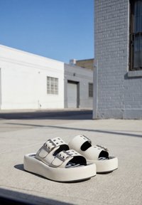 Sandales à plateforme blanches avec doubles brides à boucle posées sur un pavement en béton en extérieur devant des murs de bâtiments urbains.