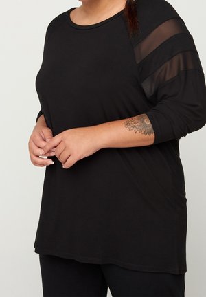 Osoba nosząca czarną bluzkę z długim rękawem z przezroczystymi pasami na ramionach, ręce lekko splecione, pokazująca kwiatowy tatuaż na przedramieniu.