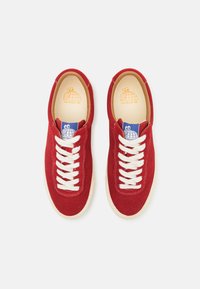 Red suede sneakers with white laces, round toe, and beige sole. Features a blue label with a logo and minimal stitching detail.