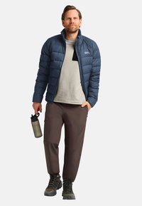 Marineblauer Pufferjacke, beiges Hemd, braune Hose, schwarze Schuhe. Hält eine graue Wasserflasche, verfügt über Reißverschlusstaschen und ein gestepptes Design.