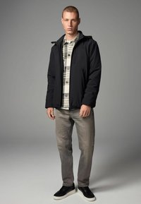 Sort letvægtsjakke med hætte, gråternet skjorte, grå jeans og sorte sneakers. Jakken har en lynlås og elastisk talje.