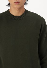 Tmavě zelený pletený svetr s žebrovaným kulatým výstřihem a texturovanou látkou. Design má dlouhé rukávy a volný střih.