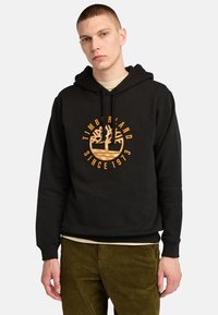 Felpa nera con cappuccio caratterizzata da un prominente logo arancione Timberland con design ad albero e testo "SINCE 1973." Realizzata in tessuto liscio con tasca a marsupio.
