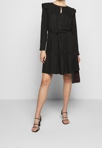 Schwarzes langärmliges Kleid mit gerafften Schultern, Schlüsselloch-Ausschnitt, gebundenem Taillenbund und gestuftem Rock. Das Model trägt schwarze Riemchen-Sandalen und hält eine dunkle Tasche.