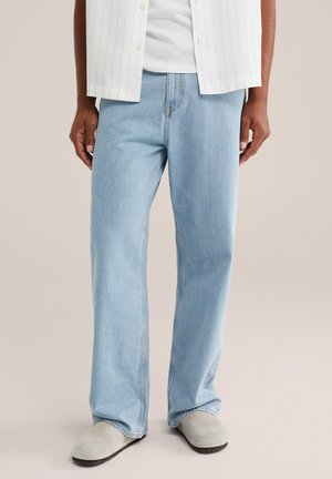 Personne portant un jean bleu clair droit, une chemise blanche et des chaussures légères à enfiler, debout sur un fond uni.