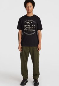 Must must-shirt valge graafika tekstiga, koos oliivroheliste cargo pükste ja mustade nööbiga saapadega.