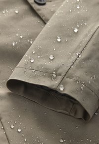 Gros plan sur la manche d'une veste imperméable beige avec des gouttelettes d'eau perlant à la surface du tissu.