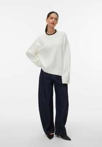 Pull blanc oversize avec poignets côtelés, associé à un jean large bleu foncé avec coutures apparentes et des escarpins noirs pointus.