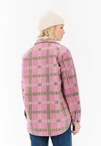 Vrouw met blond haar, die een beige gebreide muts en een roze patronenjas draagt, zijwaarts gericht tegen een witte achtergrond.